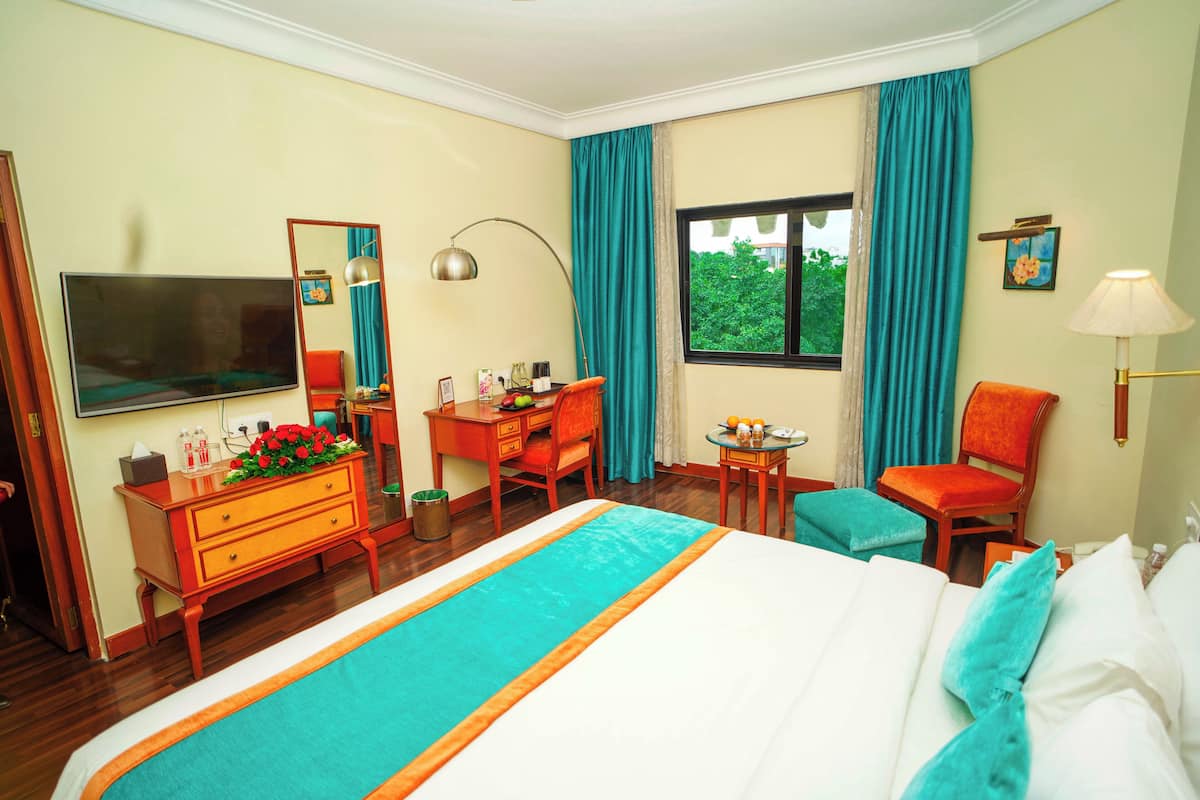Habitación de hotel con una cama grande, un escritorio con silla, televisor, ventana con cortinas y una mesita con lámpara.
