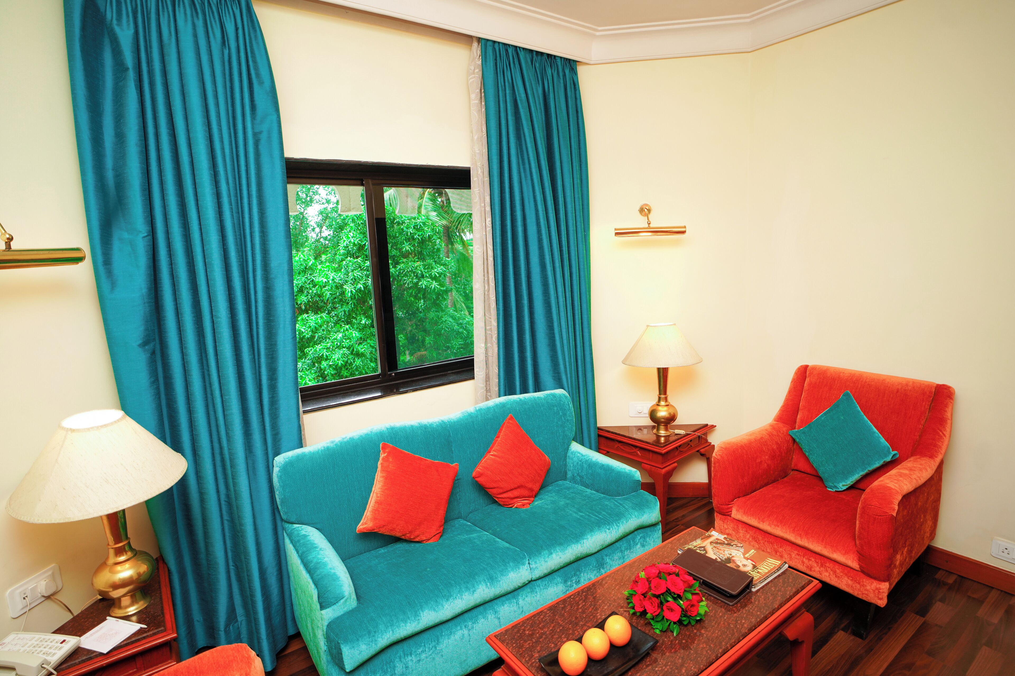 A room with a teal sofa, a red armchair, a wooden coffee table with fruits and a book, and a window with curtains.