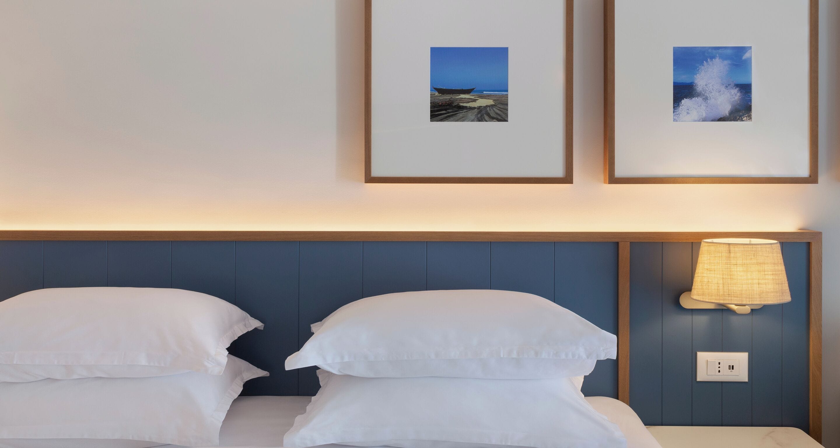 A hotel room with a bed, two pillows, a nightstand with a lamp, and two framed pictures on the wall.