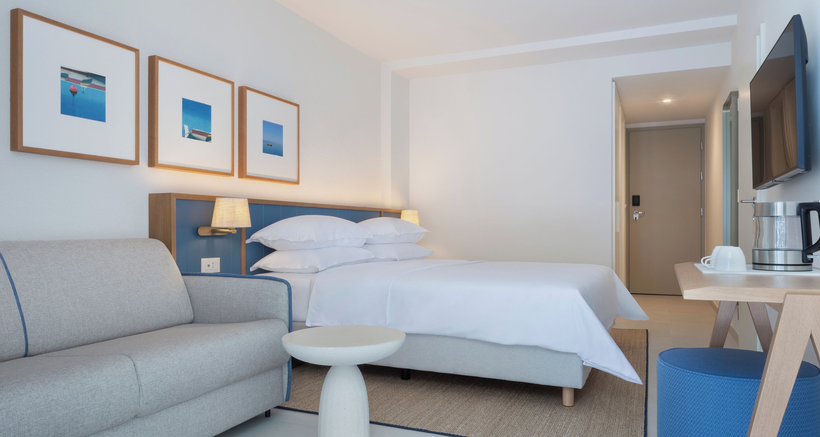 A hotel room with a bed, a sofa, a small table, a coffee maker, and framed pictures on the wall.