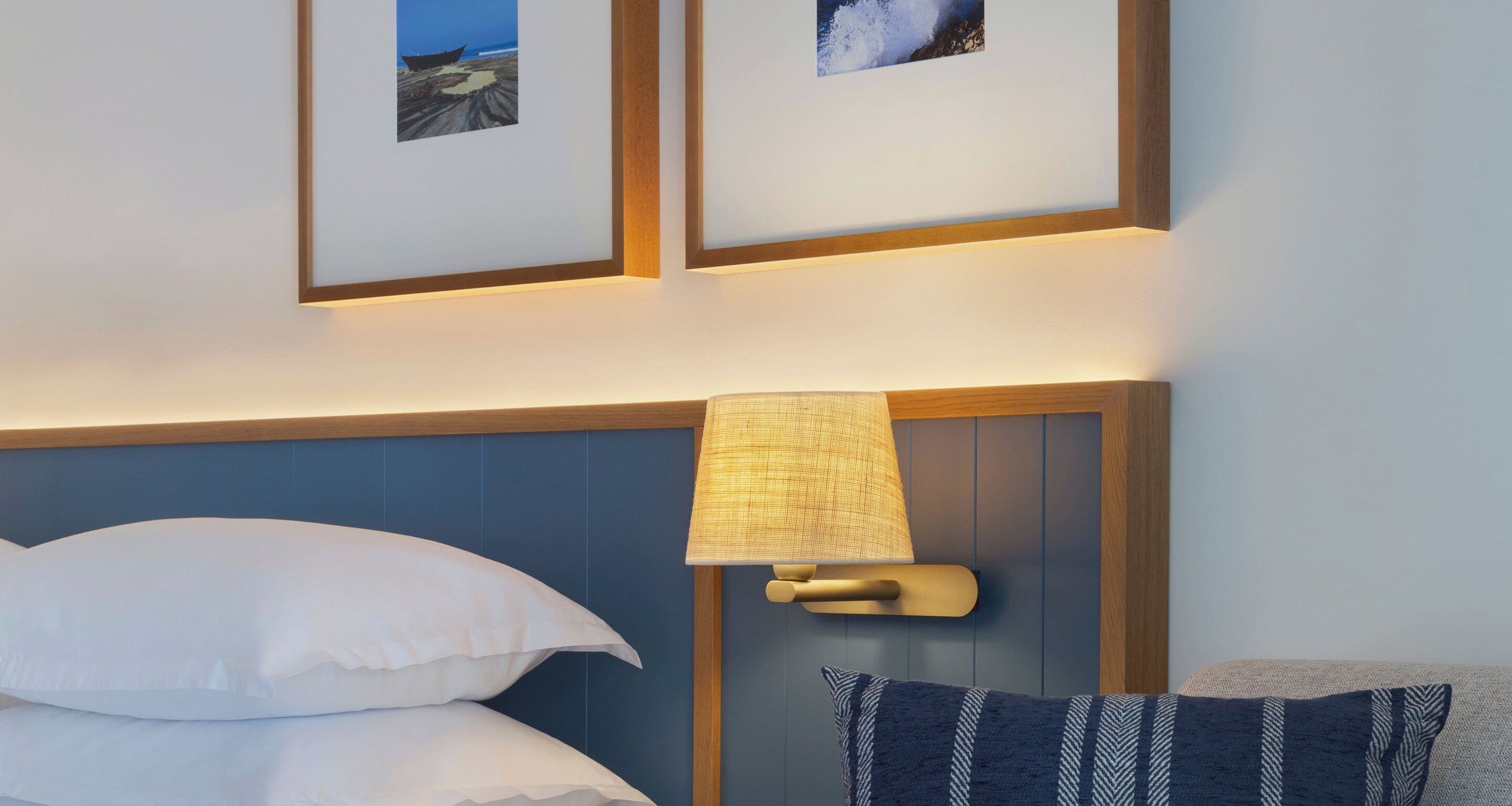 Ein ordentlich bezogenes Bett mit weißen Kissen und einem blauen Kopfteil. An der Wand hängt eine Lampe mit beigem Lampenschirm und Holzrahmen. Zwei gerahmte Bilder über dem Bett zeigen Landschaften.
