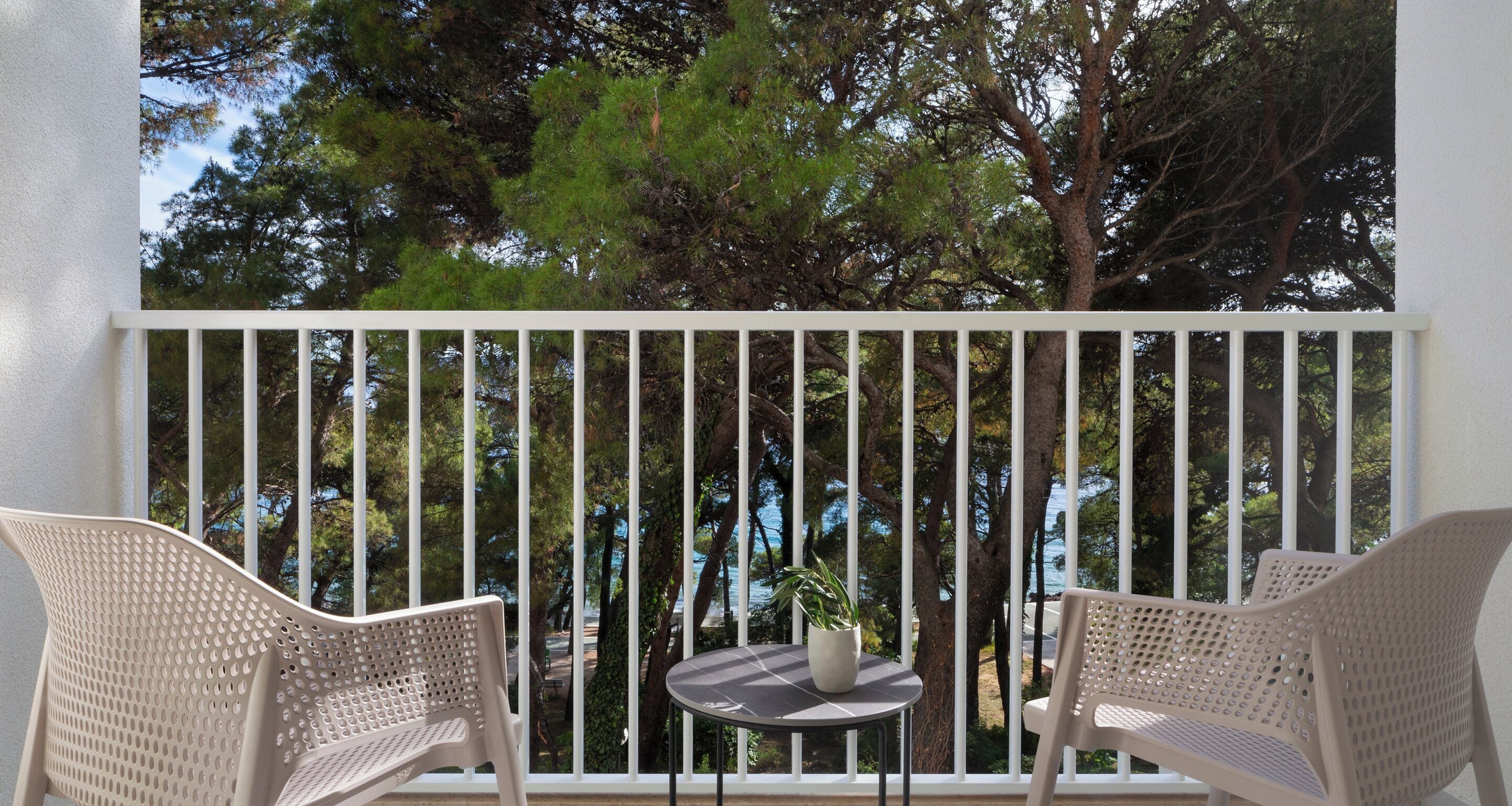 Un balcon avec deux fauteuils en osier et une petite table ronde, donnant sur une zone côtière bordée d’arbres et offrant une vue sur la mer.