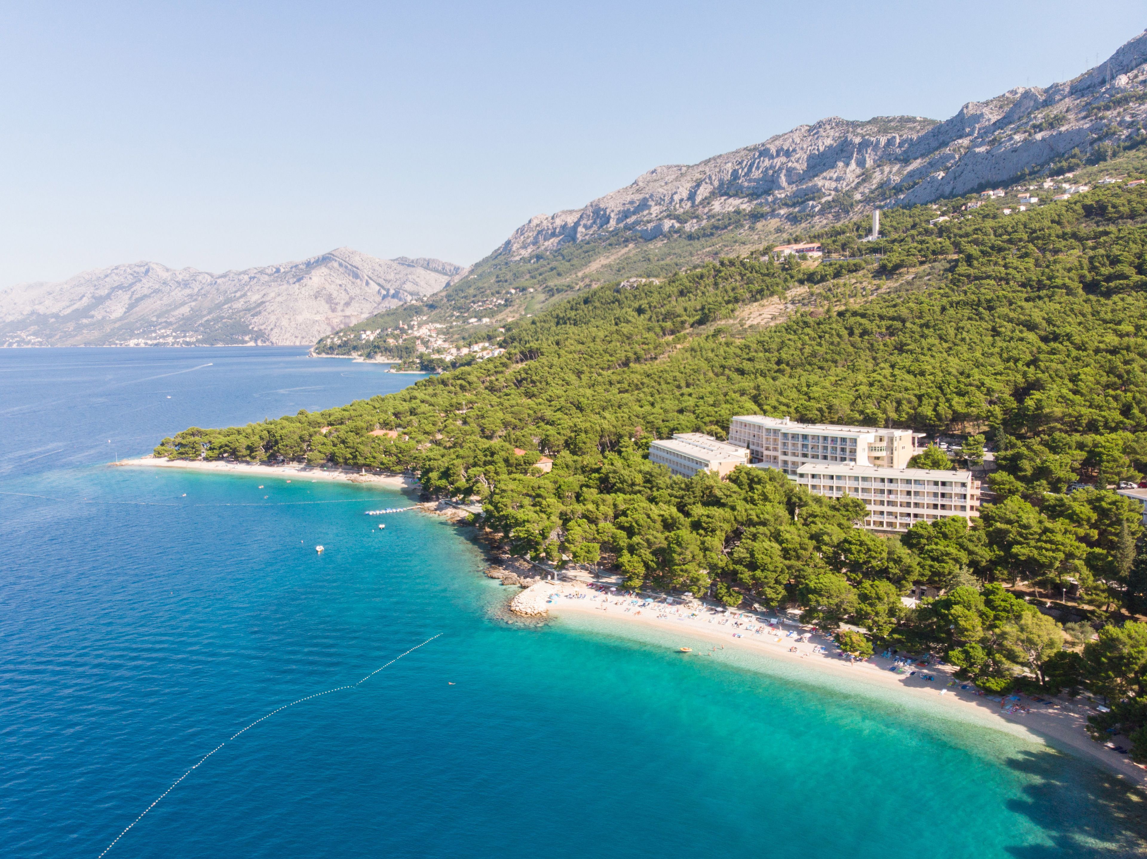 A coastal hotel with a beach, surrounded by mountains and clear blue waters.