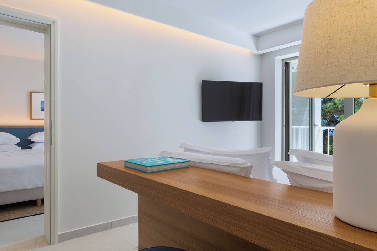 Una habitación de hotel moderna con un escritorio de madera, un televisor de pantalla plana montado en la pared y un amplio ventanal que proporciona luz natural.