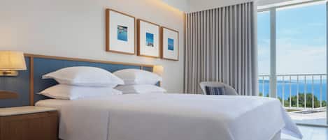 A hotel room with a large bed, a chair, a nightstand, and a view of the sea through large windows.