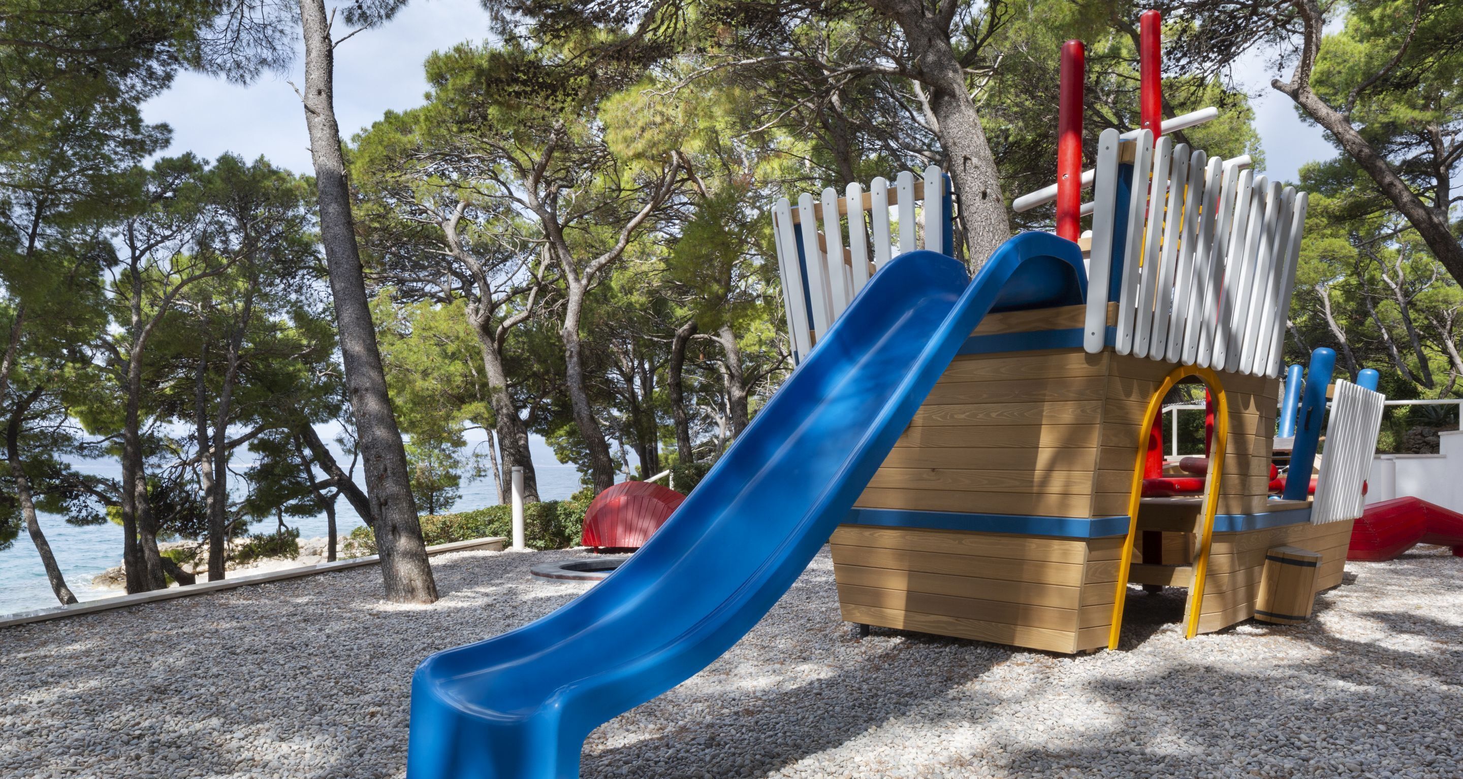 Un parque infantil con un tobogán azul y zona de escalada, rodeado de árboles y una masa de agua.