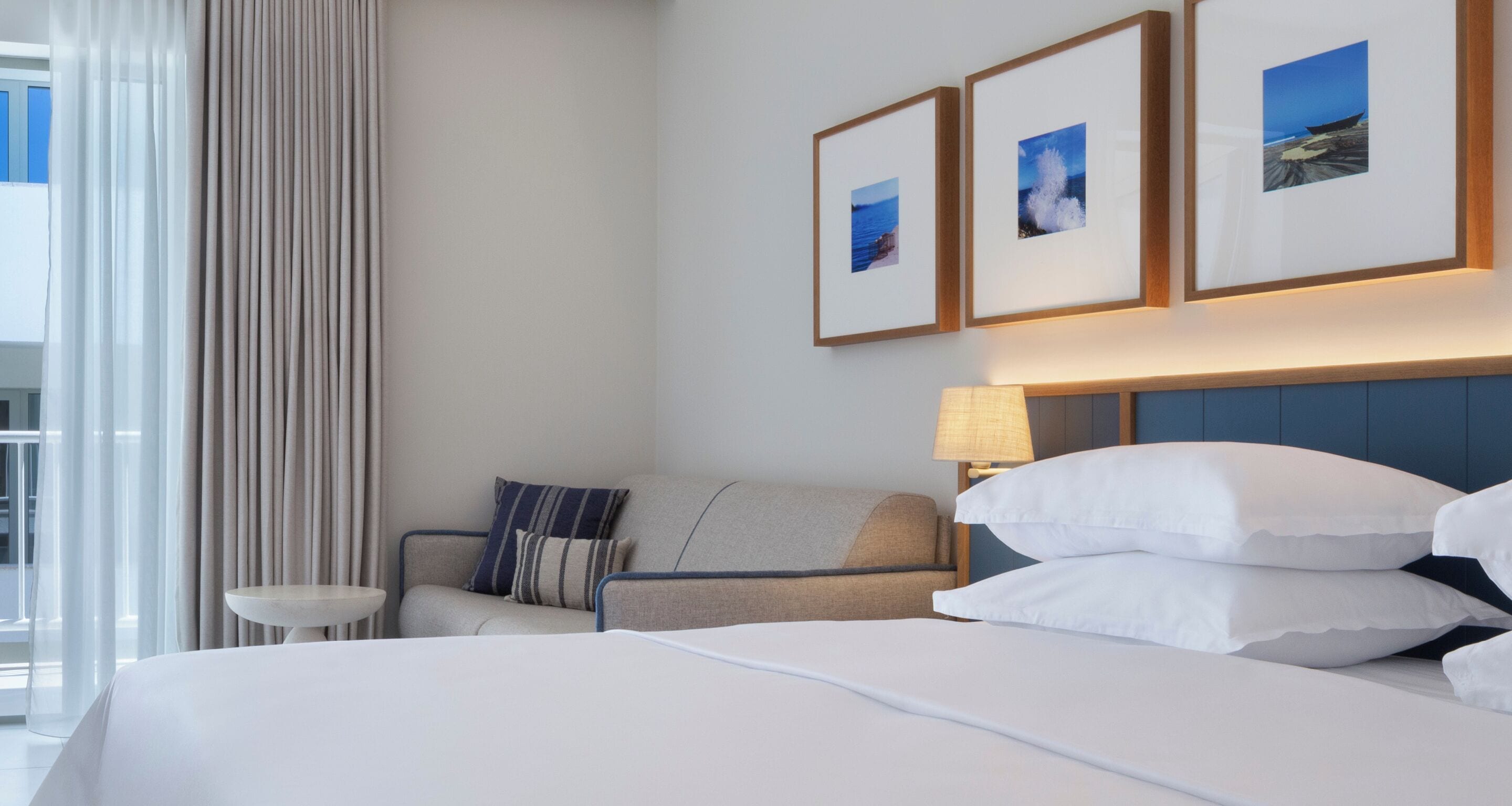 A hotel room with a bed, a sofa, a small table, and a lamp. There are three framed pictures on the wall.