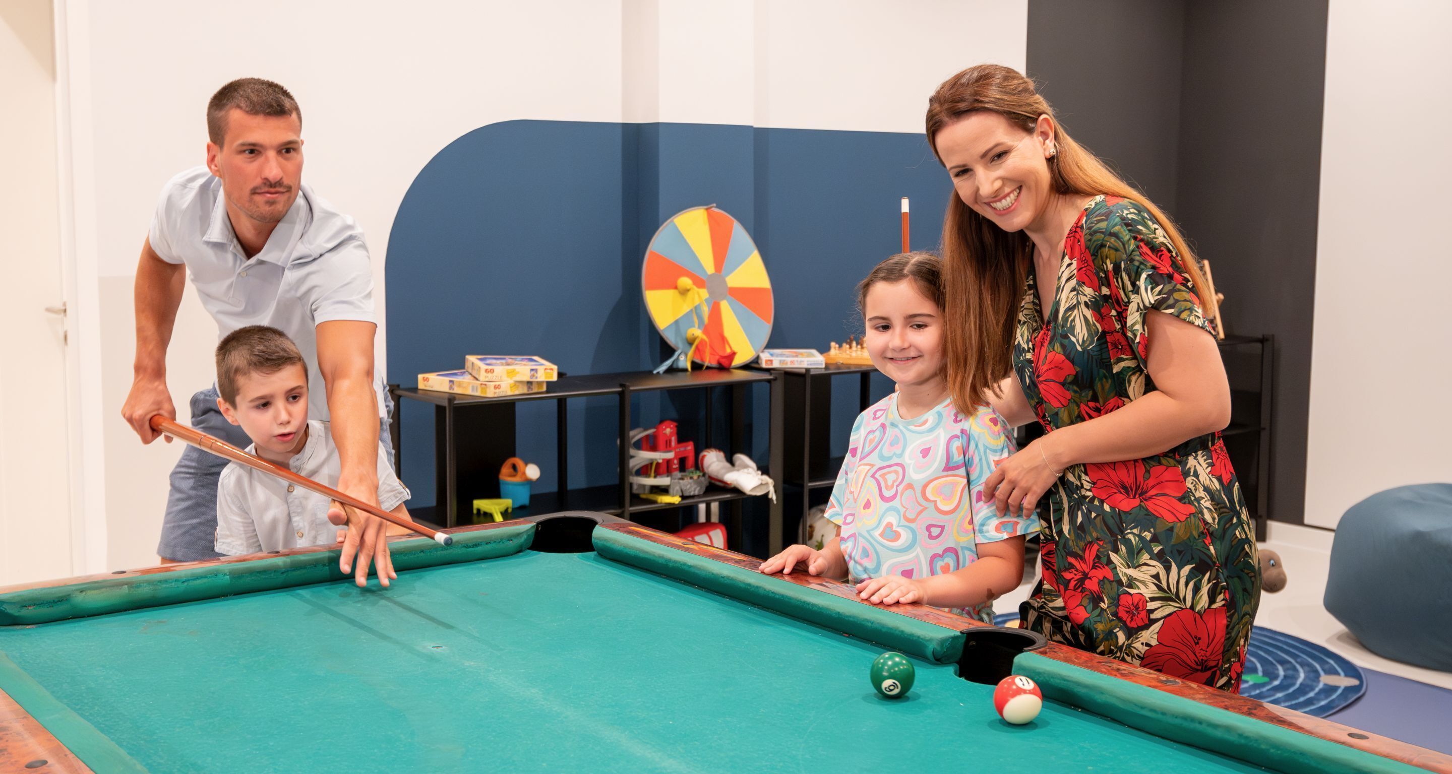 A pool table with balls, a dartboard, and a shelf with toys and shoes in a room with blue walls.