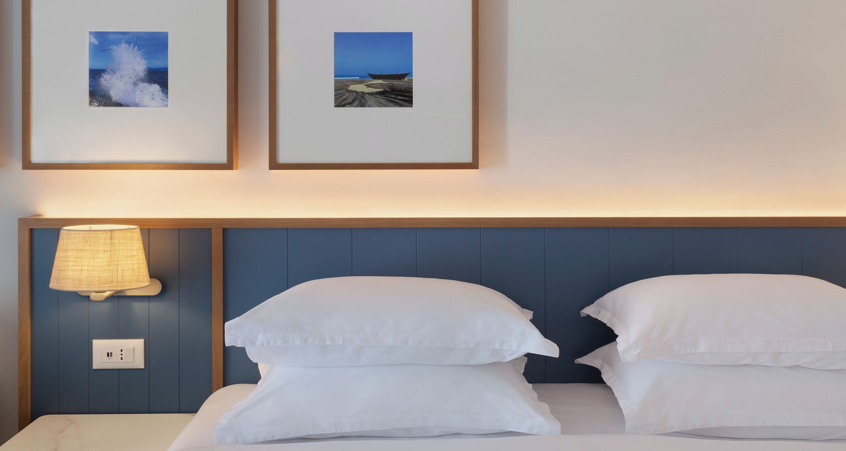 Quarto de hotel com uma cama, duas almofadas, uma luminária fixada na parede e duas imagens emolduradas com uma paisagem de praia.