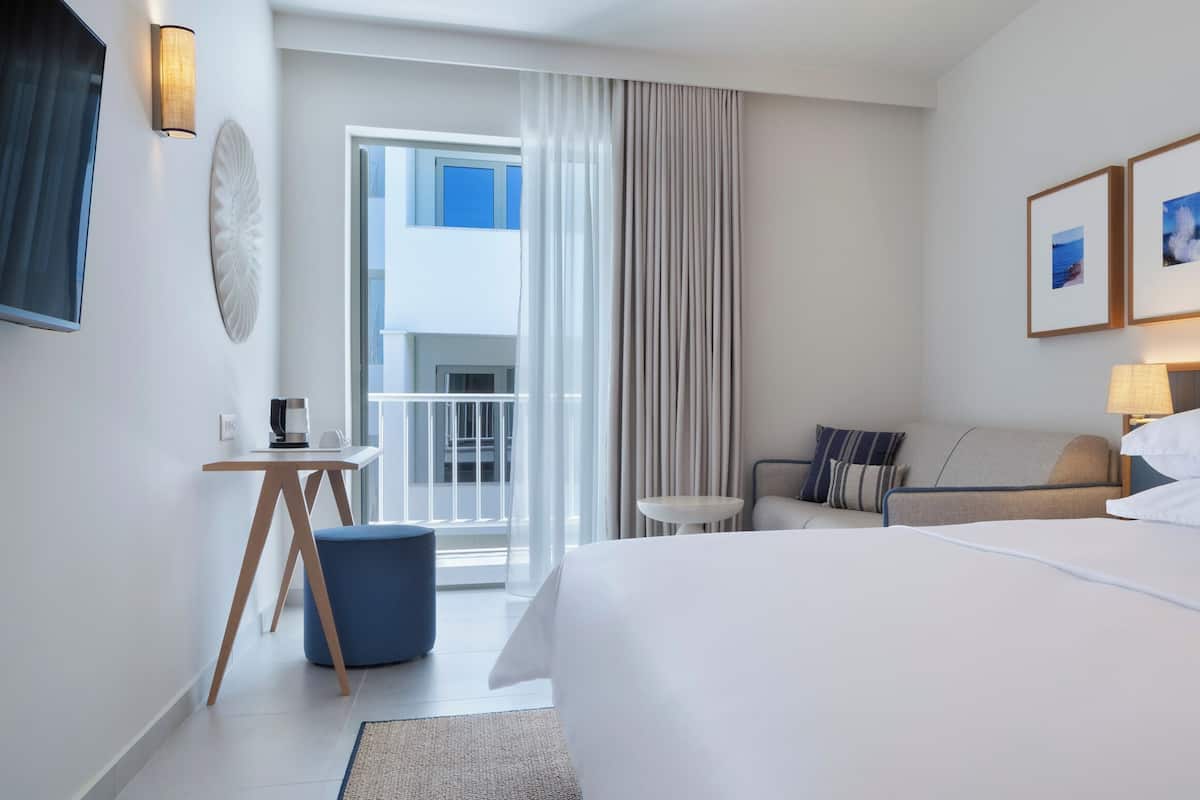 Habitación de hotel con una cama grande, un escritorio con cafetera, una mesita redonda pequeña, un sofá y vistas al balcón.