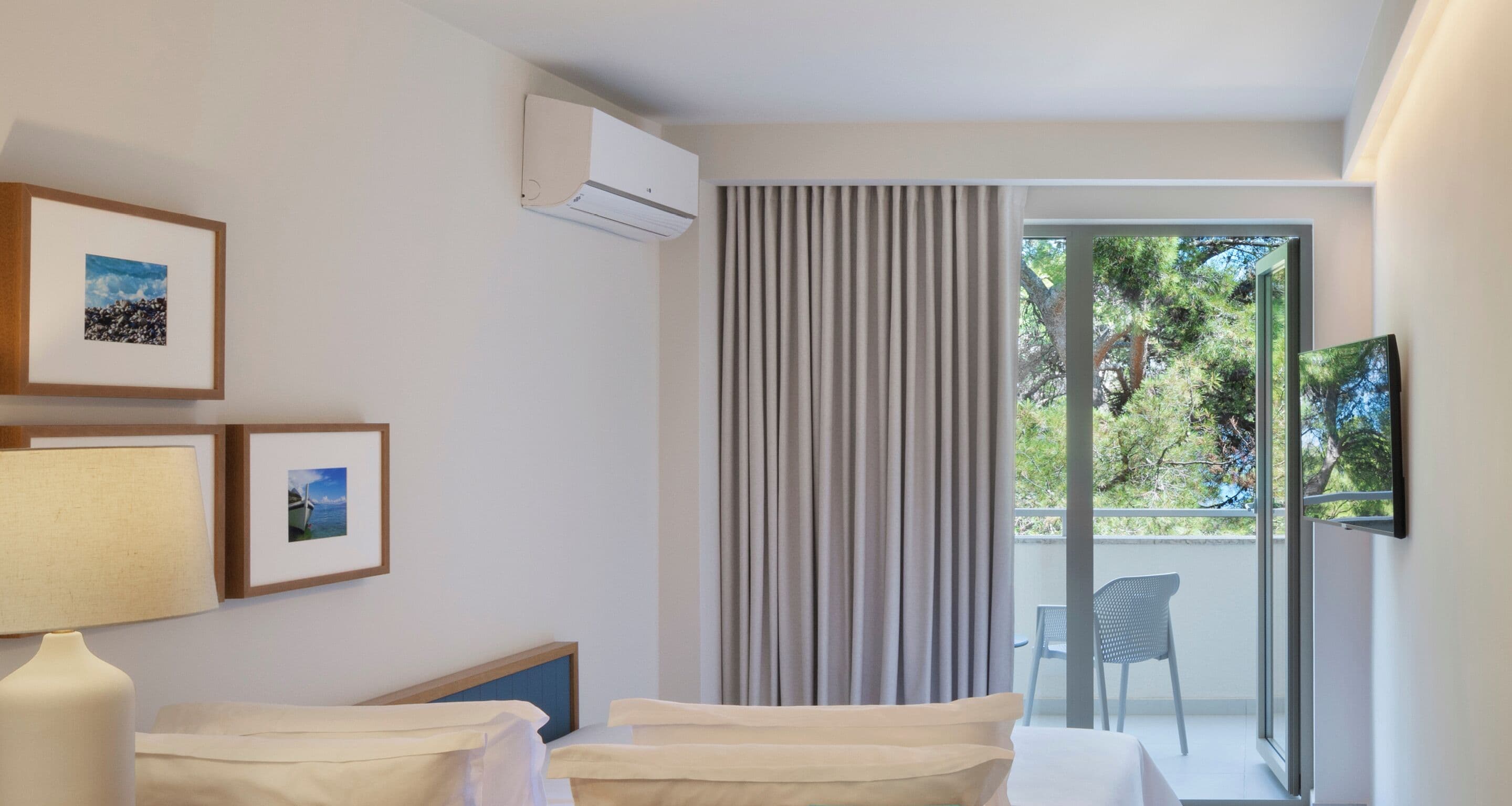 Habitación de hotel con cama, escritorio, silla, aire acondicionado y balcón con vista a árboles.