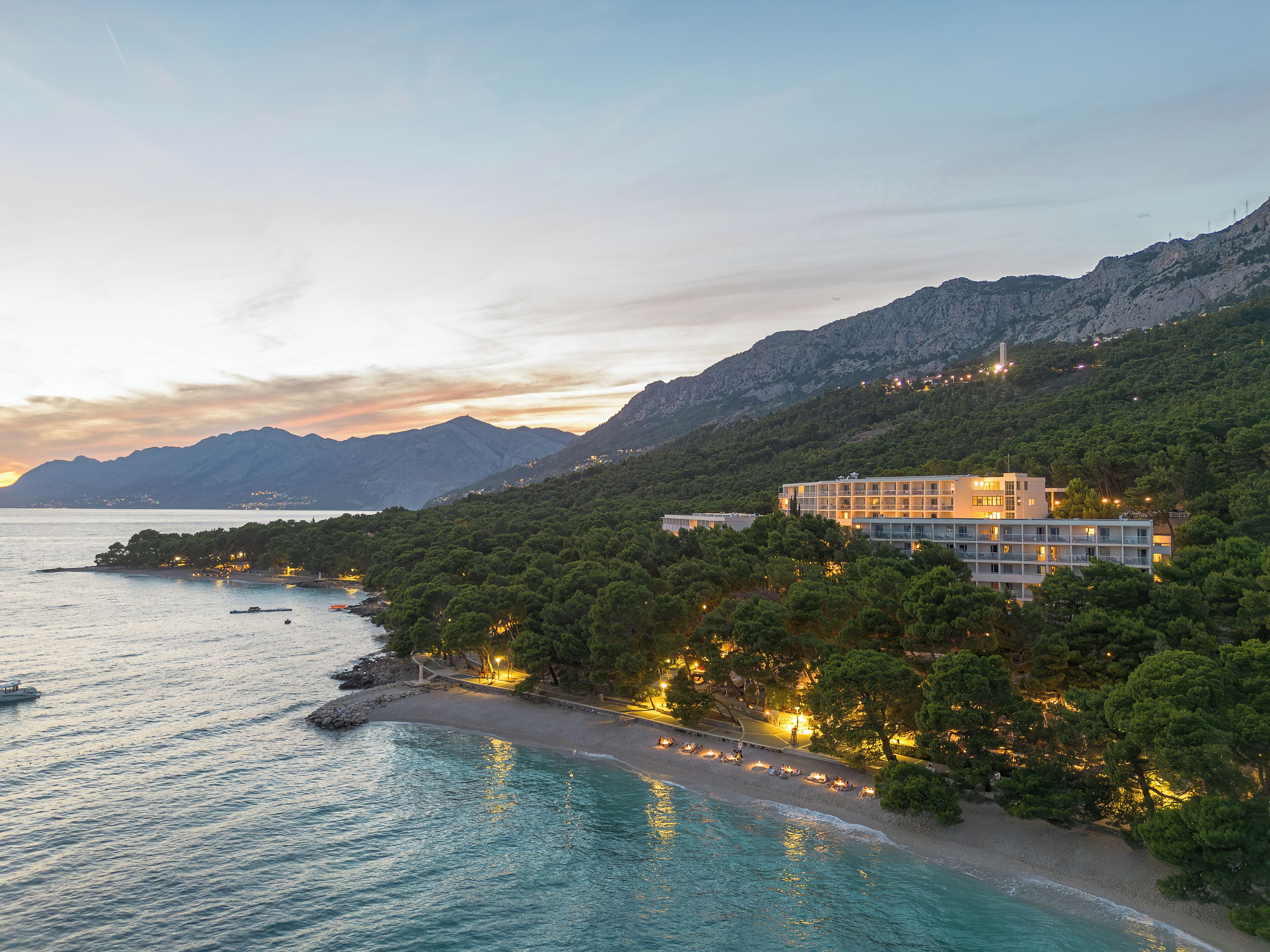 A coastal hotel with a view of the sea and mountains, surrounded by greenery.