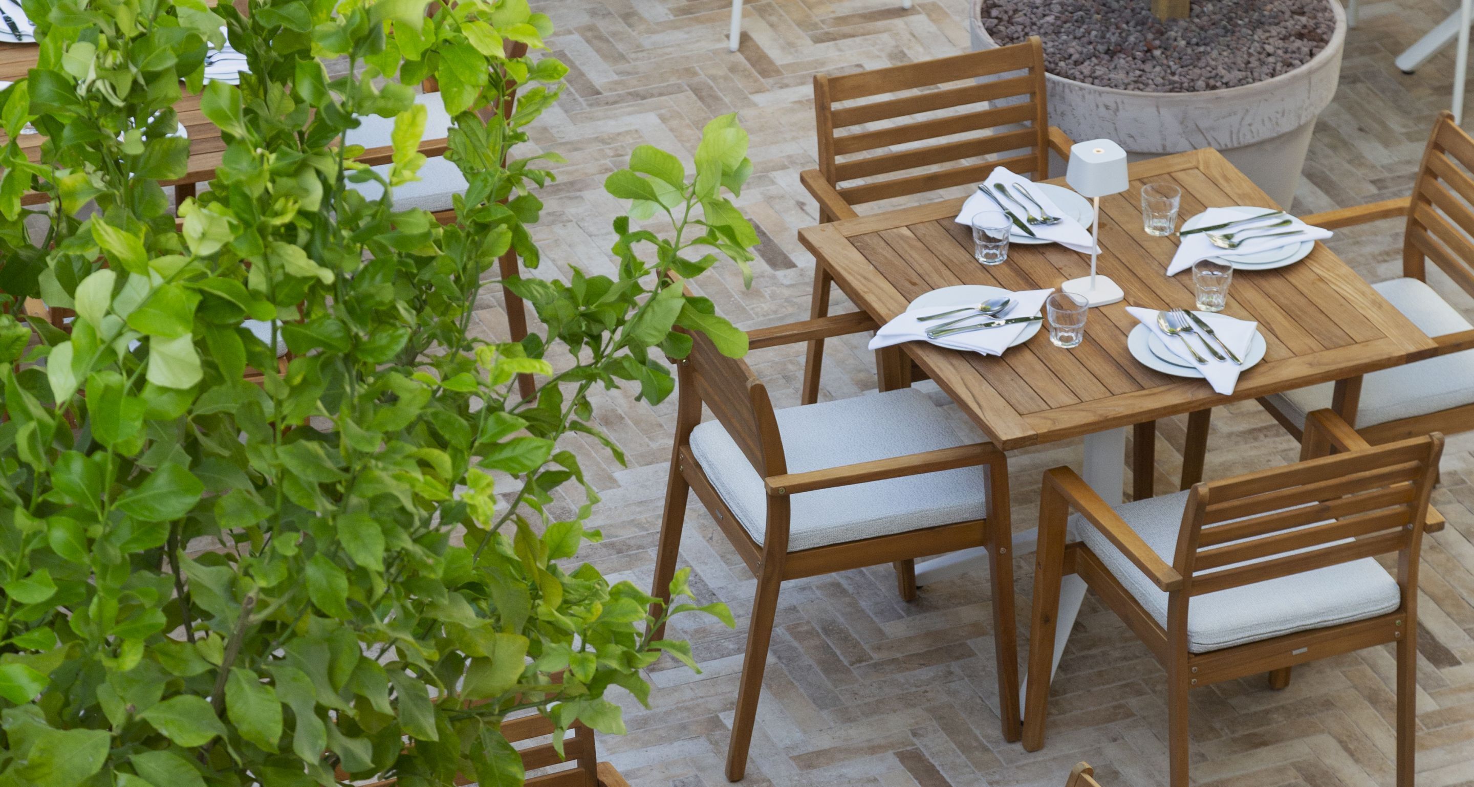 Een houten eettafel, gedekt met witte borden, bestek en glazen, omringd door houten stoelen met kussentjes.