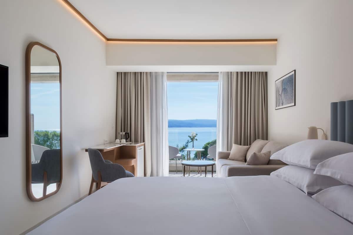 A hotel room with a large bed, a desk, a chair, a sofa, and a view of the sea.