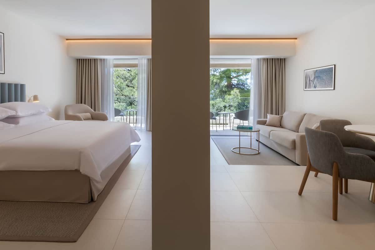 A modern hotel room with a large bed, a sofa, a dining table, and a balcony with a view of trees.