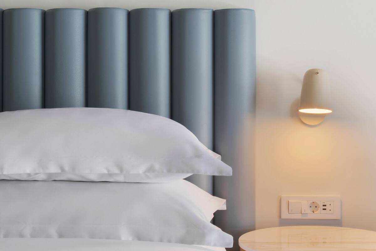 A bed with white pillows and a headboard with vertical panels, a bedside table, and a wall-mounted light fixture.