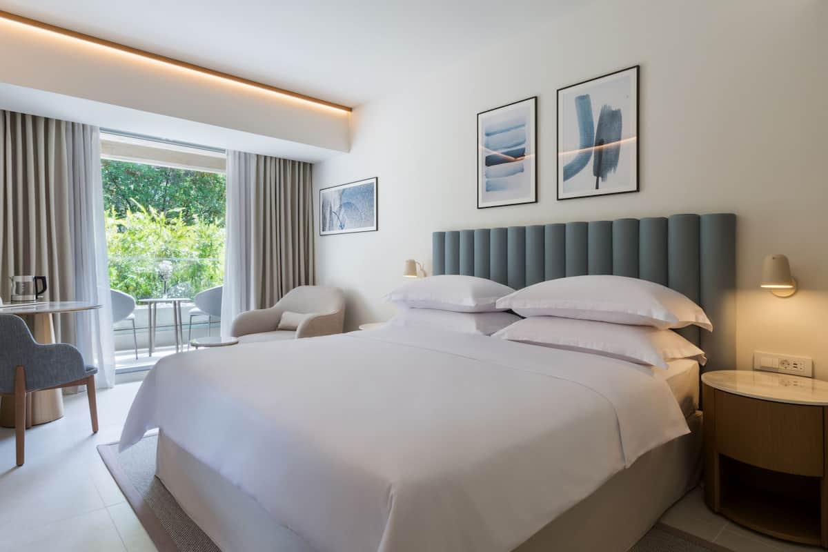 A modern hotel room with a large bed, a desk with a chair, a small table, and a view of greenery outside.