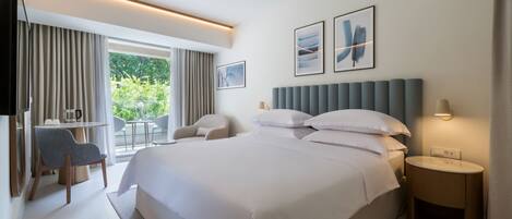 A modern hotel room with a large bed, a desk with a chair, a small table, and a view of greenery outside.