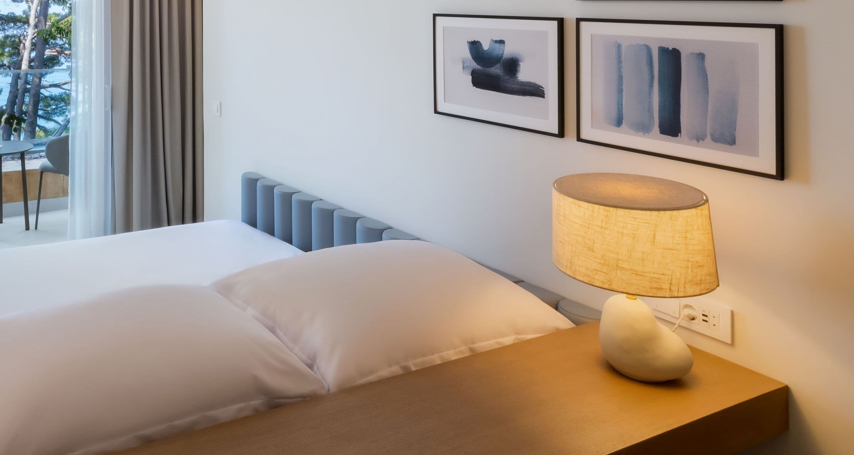 A hotel room with a bed, a bedside lamp, and two framed artworks on the wall.