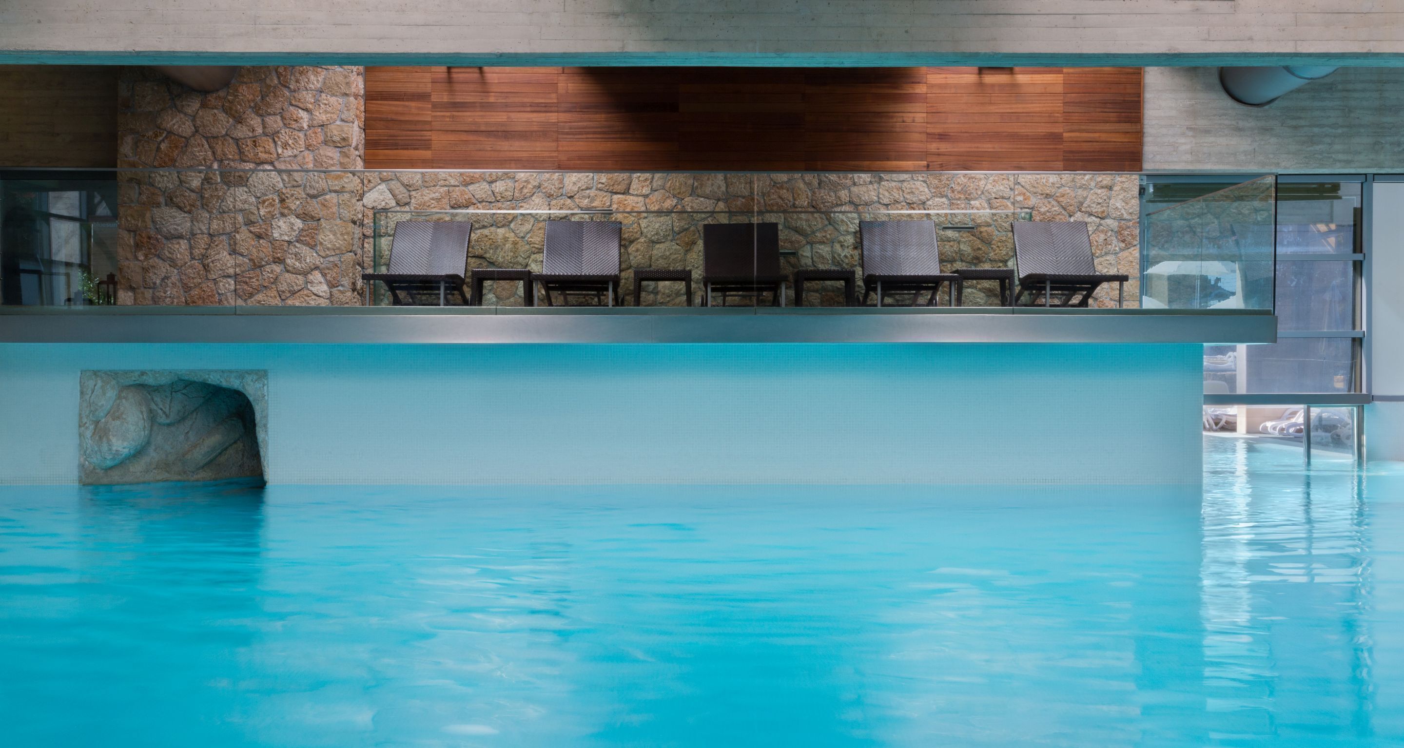 An indoor swimming pool with stone walls and wooden paneling, surrounded by six lounge chairs.