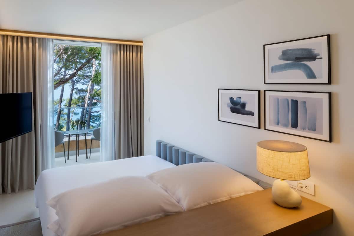 A hotel room with a bed, a TV mounted on the wall, a table with a lamp, and a balcony with a view of trees and a railing.