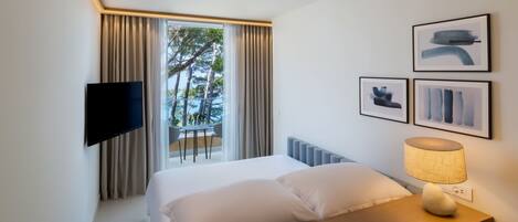 A hotel room with a bed, a TV mounted on the wall, a table with a lamp, and a balcony with a view of trees and a railing.