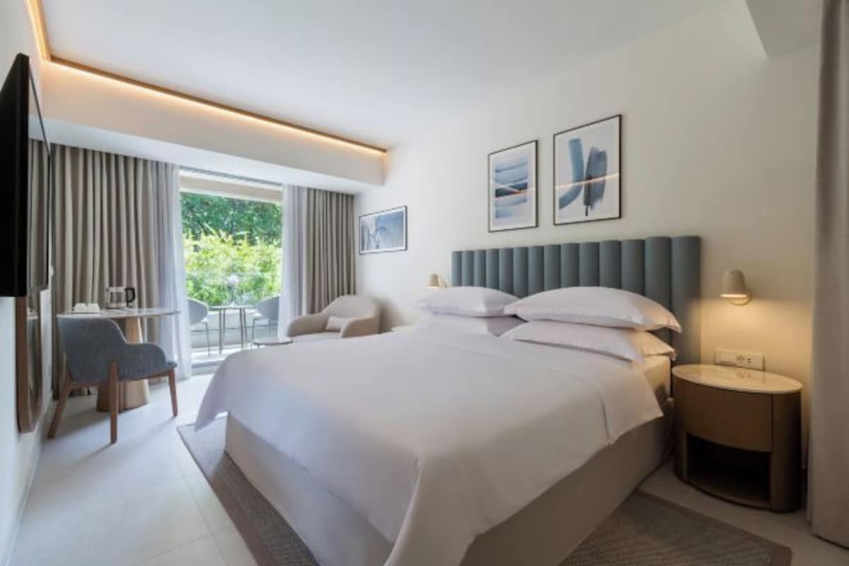 A modern hotel room with a large bed, a desk, a chair, and a view of the outdoors.