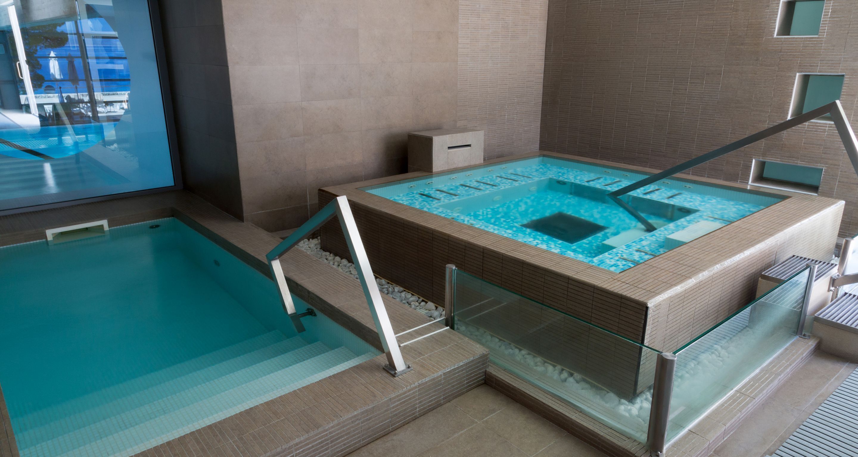 Une piscine intérieure moderne, dotée de rampes en verre et d’un espace piscine plus petit et séparé.