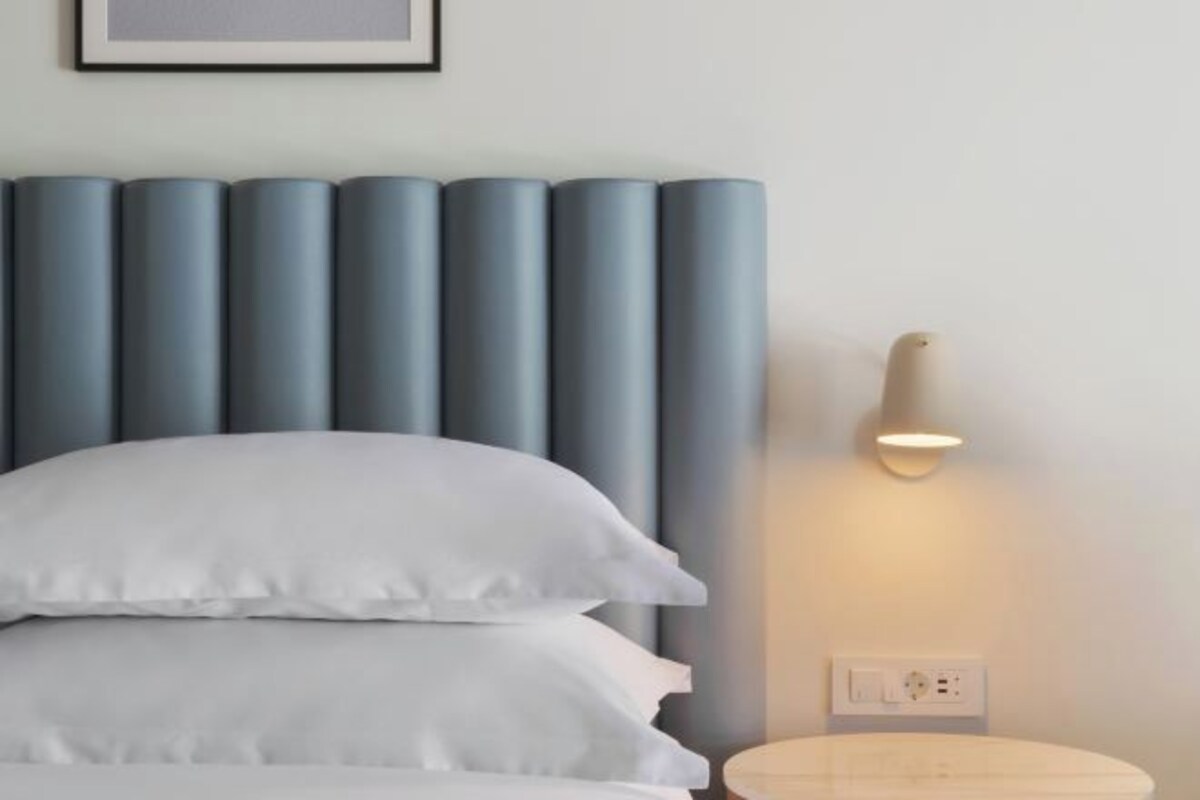 A bed with white bedding, a wall-mounted light fixture, and a power outlet on the wall.