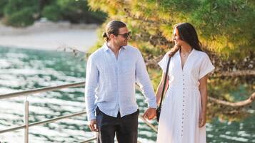 A couple holding hands on a bridge with a body of water and greenery in the background.