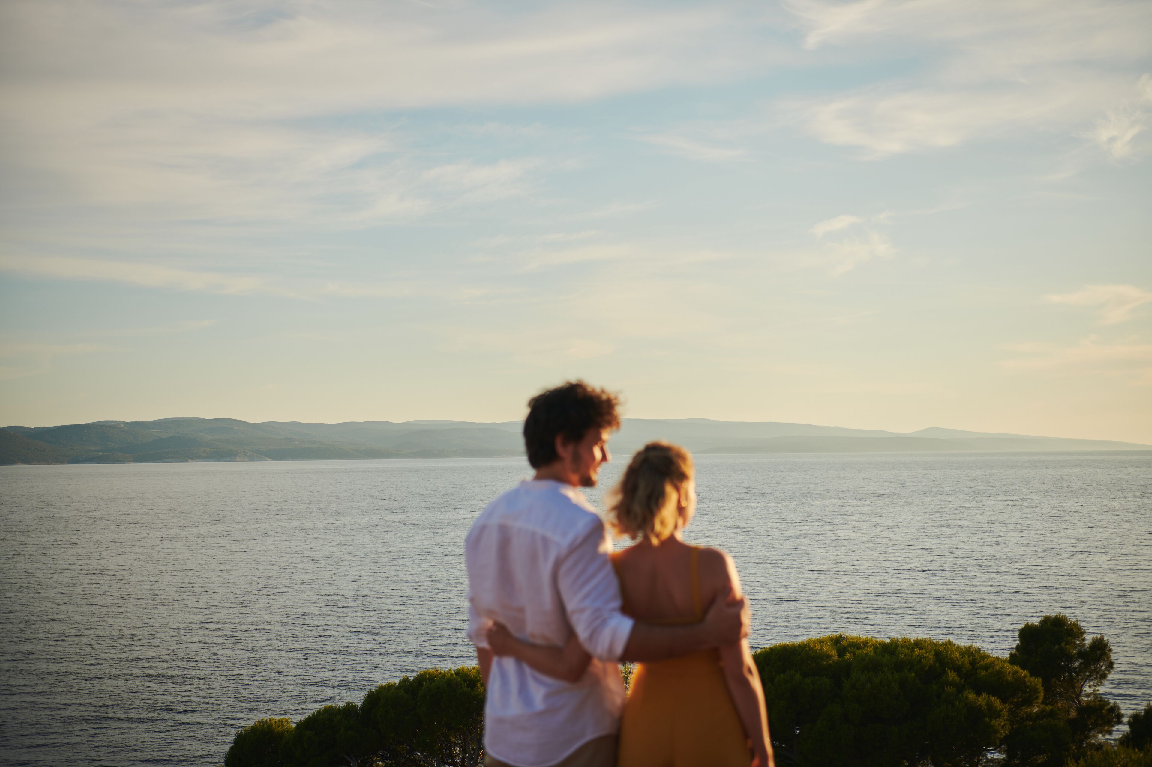 A couple standing by a body of water with a view of distant hills.