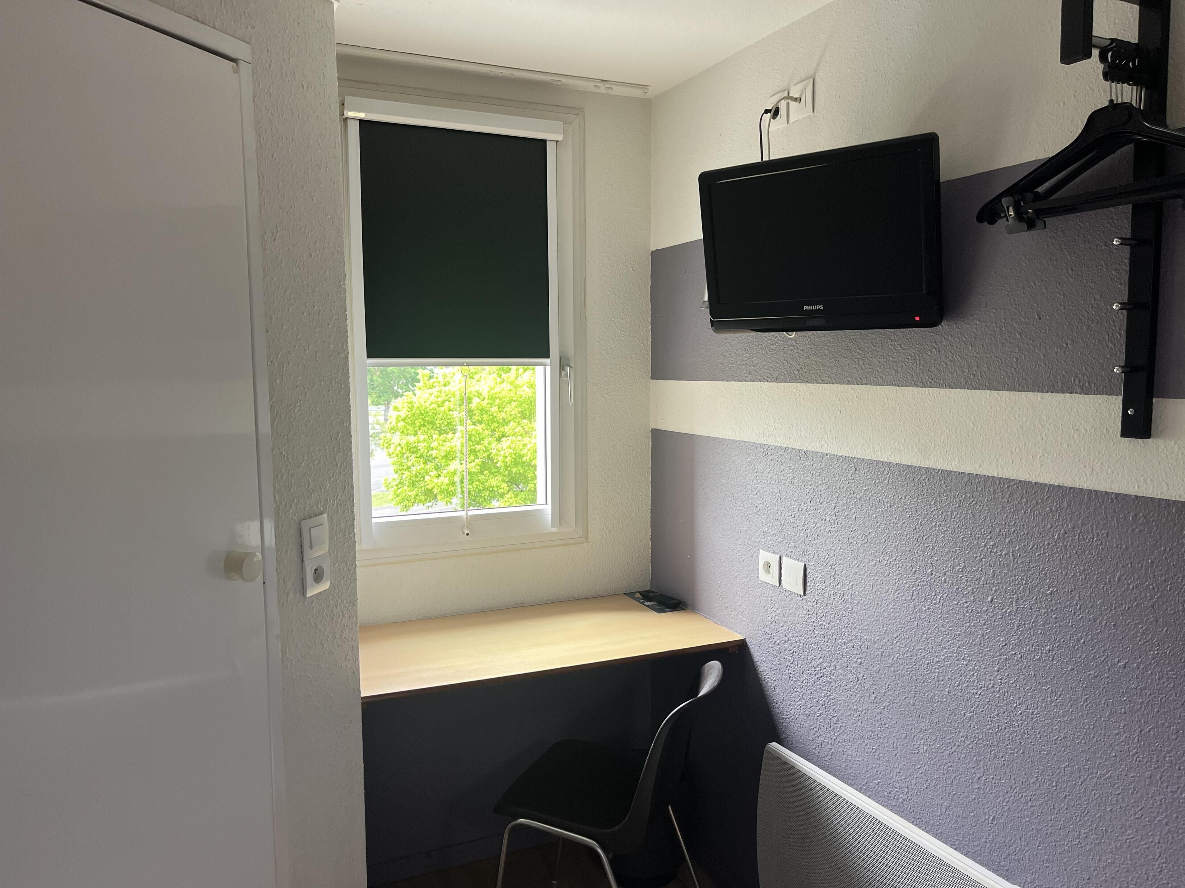 Een kamer met een raam, een wandtelevisie, een bureau en een stoel.