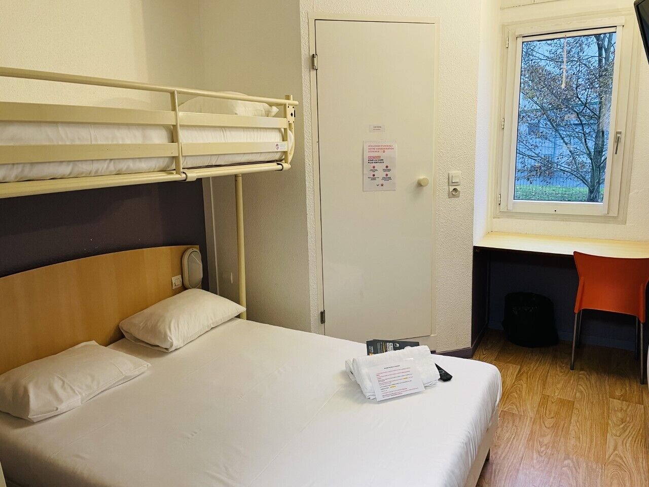 Een hotelkamer met een stapelbed, een bureau met een stoel en een raam met uitzicht op bomen.