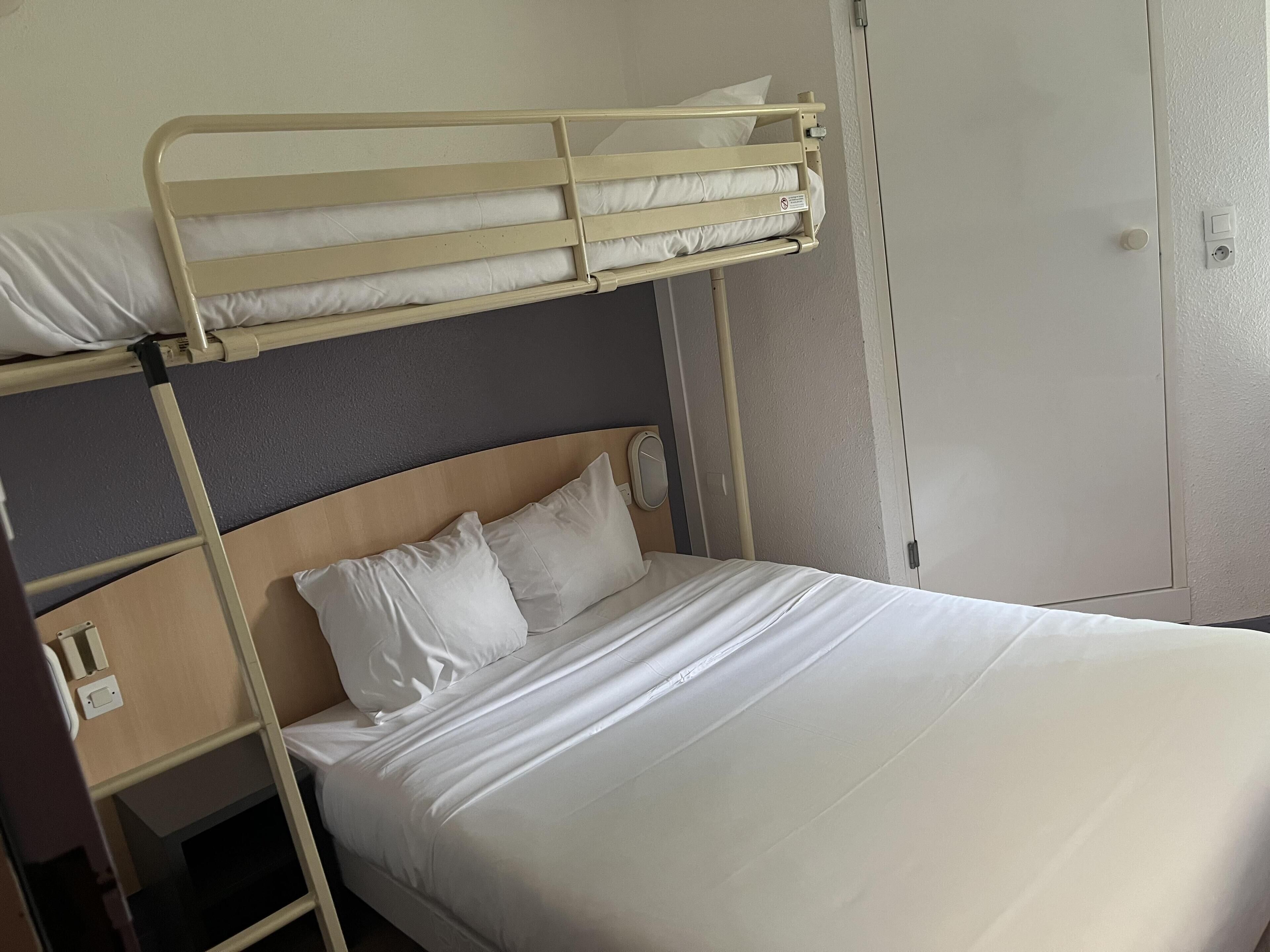 Una cama litera con cabecera de madera, una cama individual debajo y una puerta blanca a la derecha.