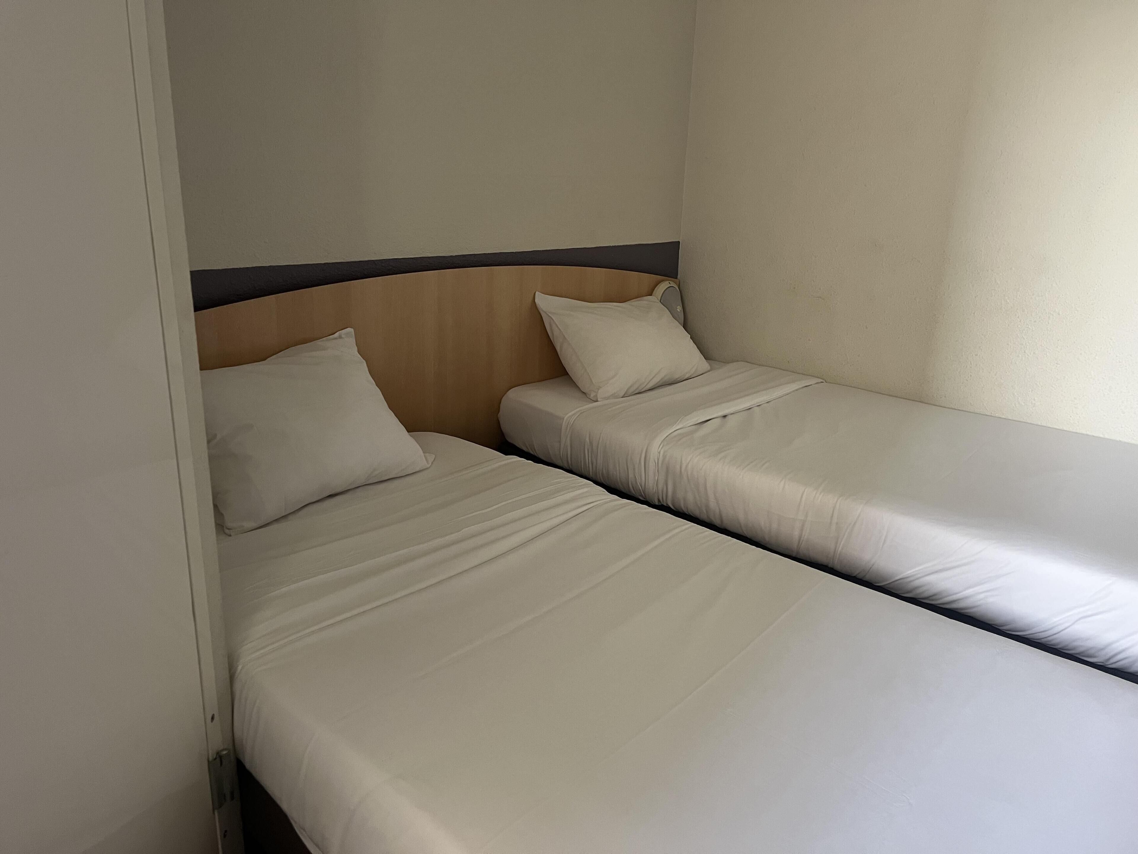 Een kleine hotelkamer met één ruimte, twee enkele bedden, een houten hoofdbord en een effen muur.