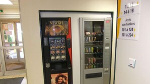 A vending machine with Nescafe coffee pods and a drink dispenser, located in a hallway with a tiled floor and a door to a room.