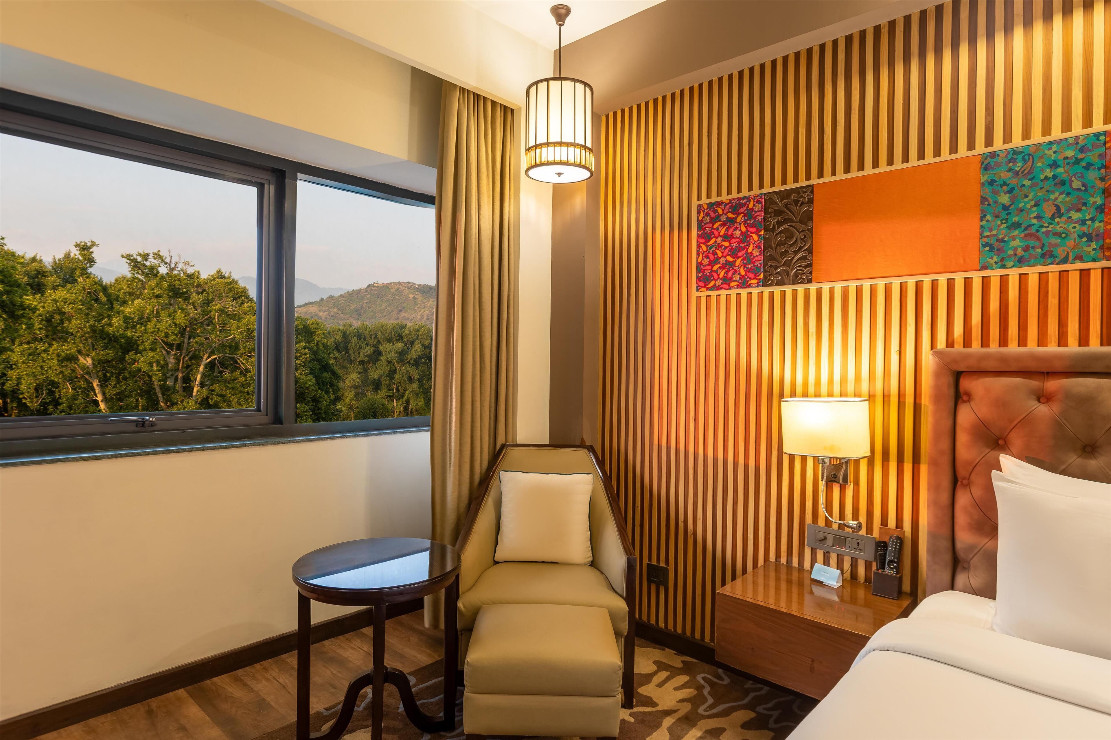 A hotel room with a large window, a chair, a bed, and a nightstand with a lamp.