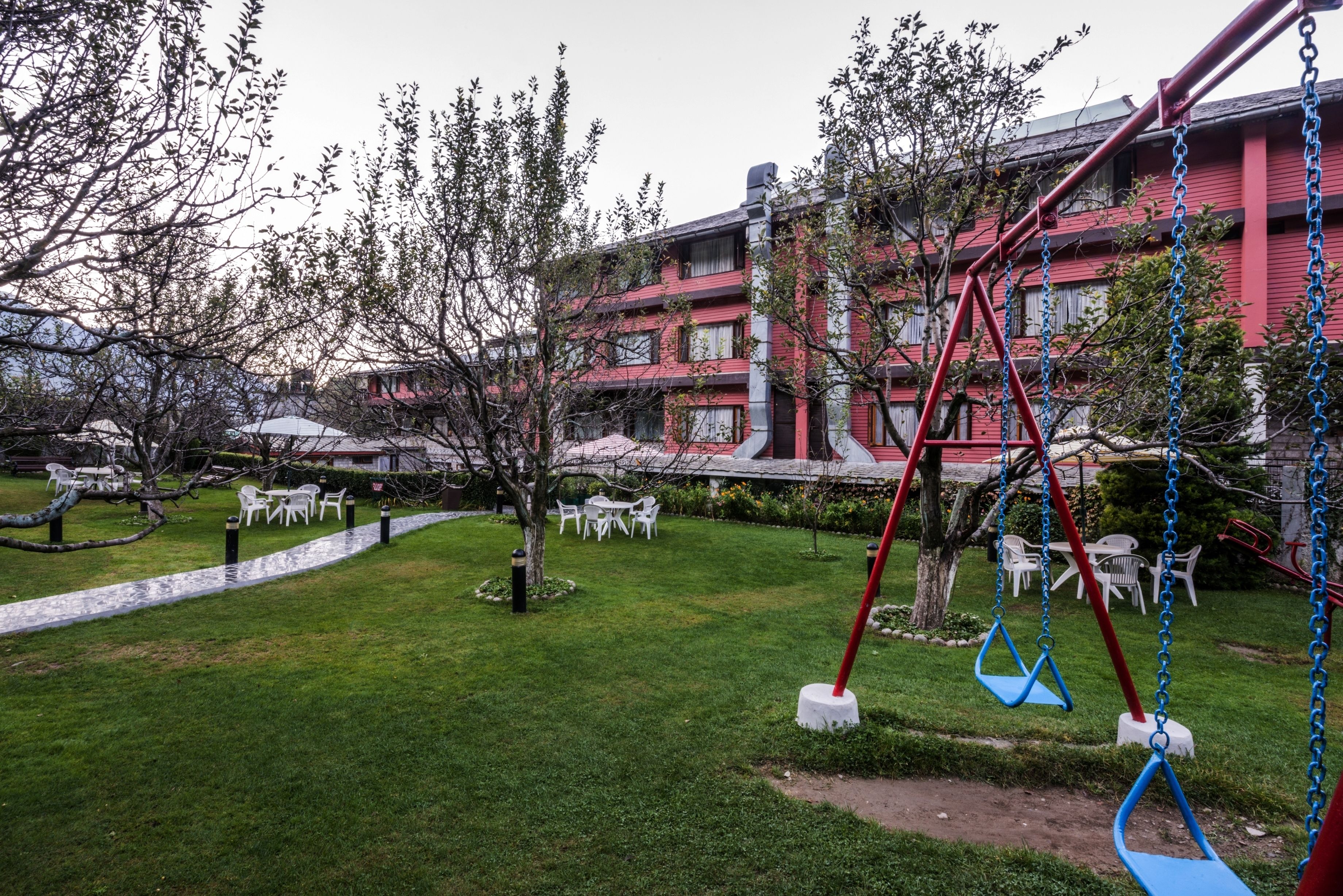 A courtyard with a swing set, white plastic chairs, and a red building in the background.