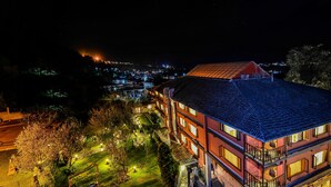 A multi-story building with a red facade and a tiled roof, featuring balconies and a well-lit garden area. - Honeymoon Inn (Manali)