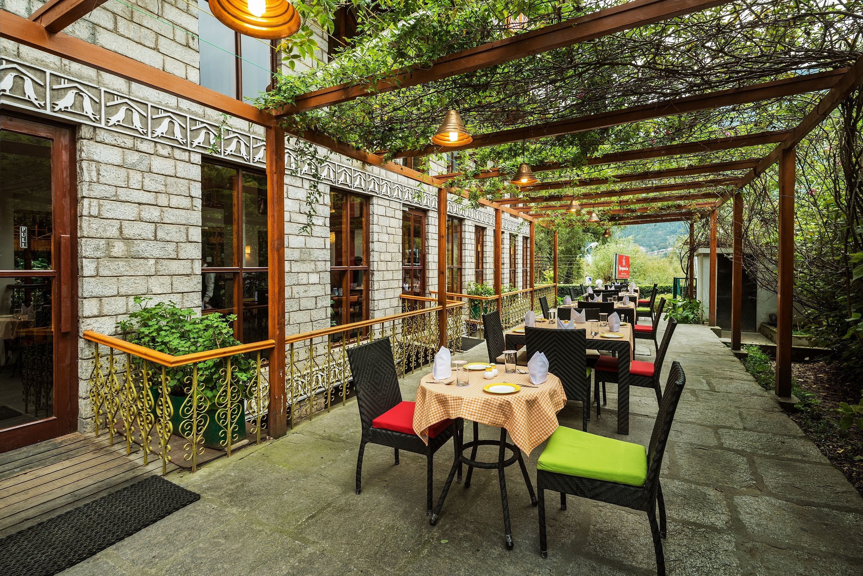 An outdoor dining area with tables and chairs, a pergola with hanging lights, and a stone building with windows and decorative railings.
