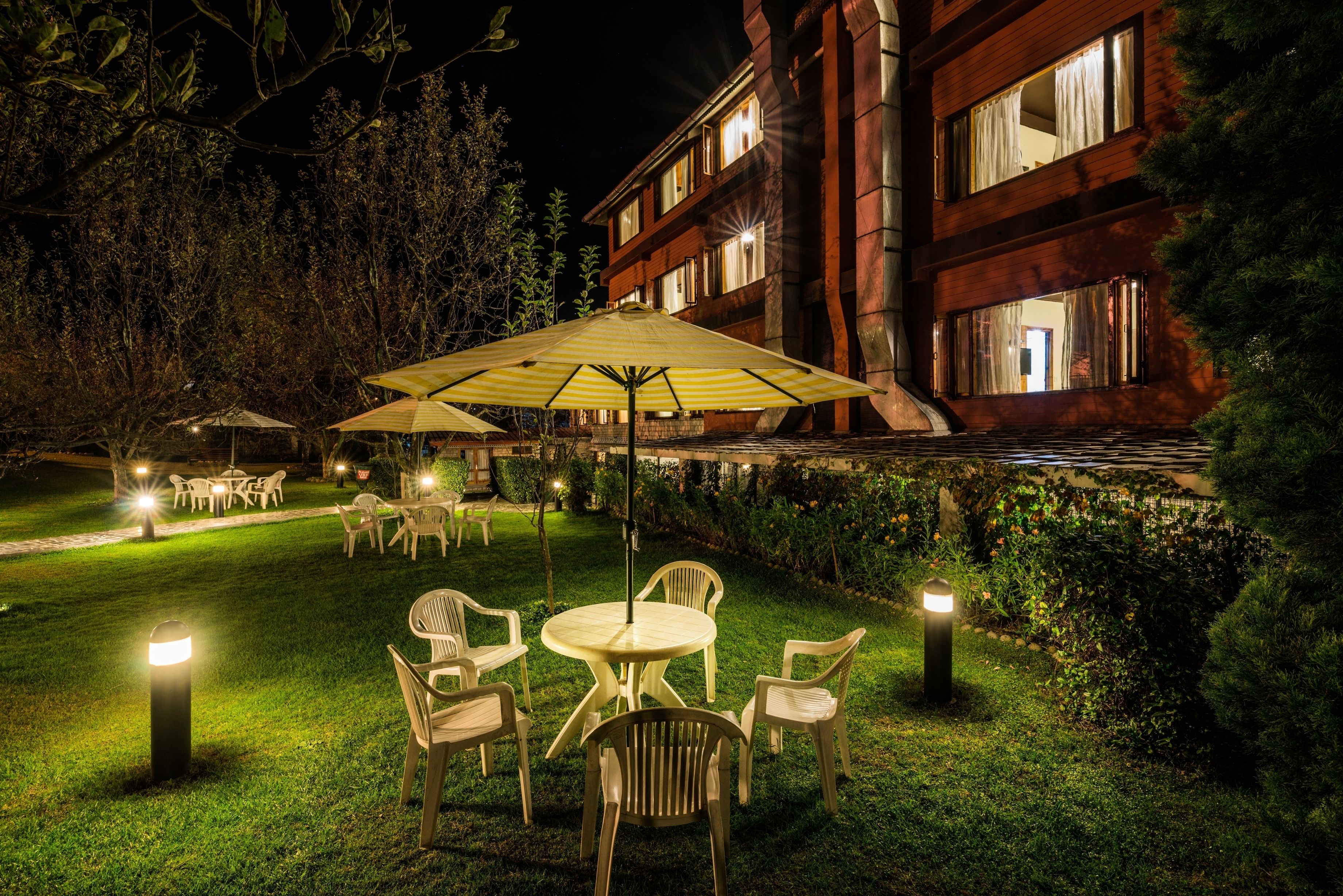 An outdoor dining area with tables and chairs, surrounded by greenery and a building with illuminated windows.