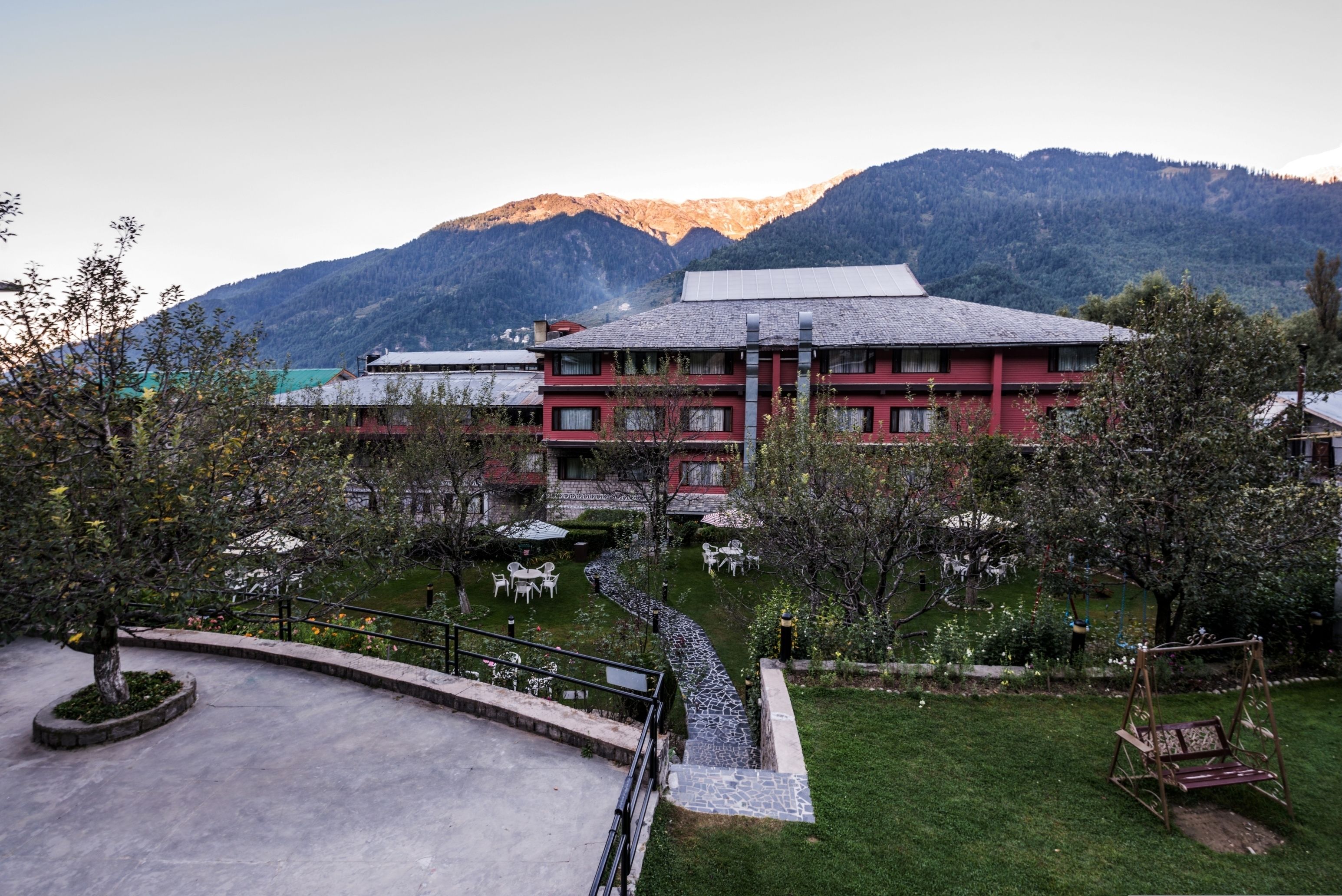 A hotel with a red facade, a garden with a pathway, and mountains in the background.