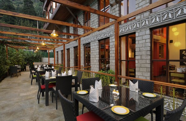 Un área de comedor al aire libre con mesas y sillas, vigas de madera y una pared de piedra.