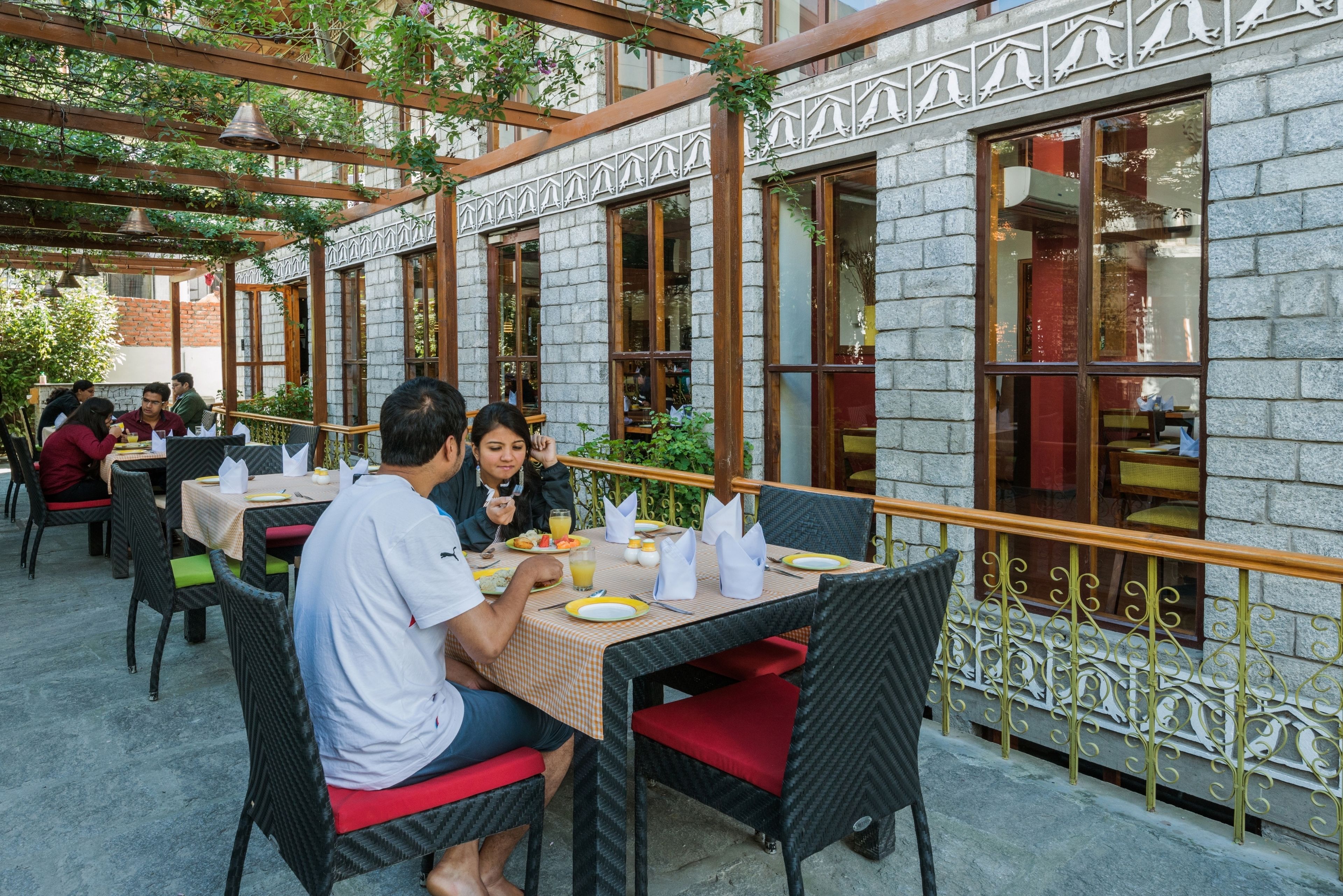 Un área de comedor al aire libre con mesas y sillas, un edificio de piedra con ventanas amplias y una barandilla de metal.
