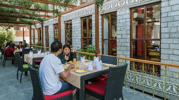 An outdoor dining area with tables and chairs, a stone building with large windows, and a metal railing.