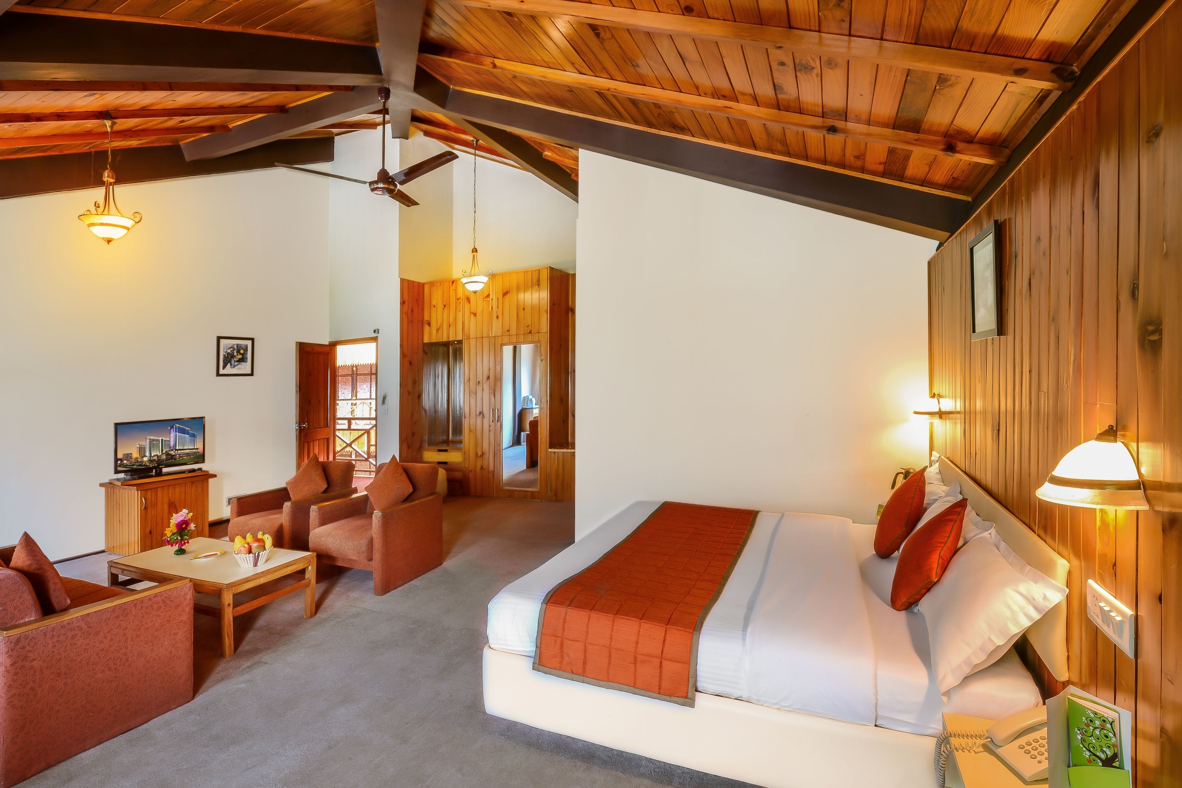 Ein Hotelzimmer mit Holzboden, einem Bett mit weißer Matratze und orangefarbener Decke, einem Holztisch mit Stuhl, einer Couch und einem Fernseher auf einem Schrank.