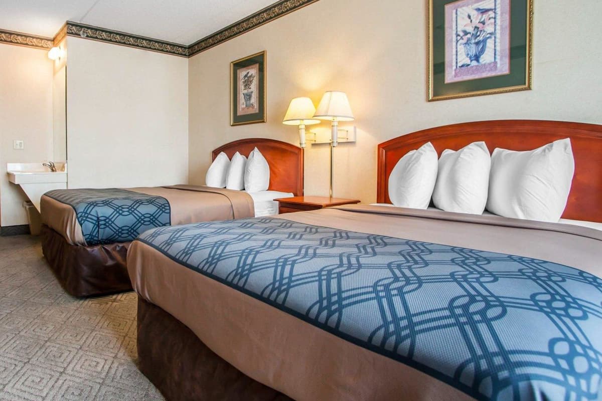 Une chambre d’hôtel avec deux lits, une table de chevet avec une lampe, un tableau encadré au mur et une salle de bain avec un lavabo.