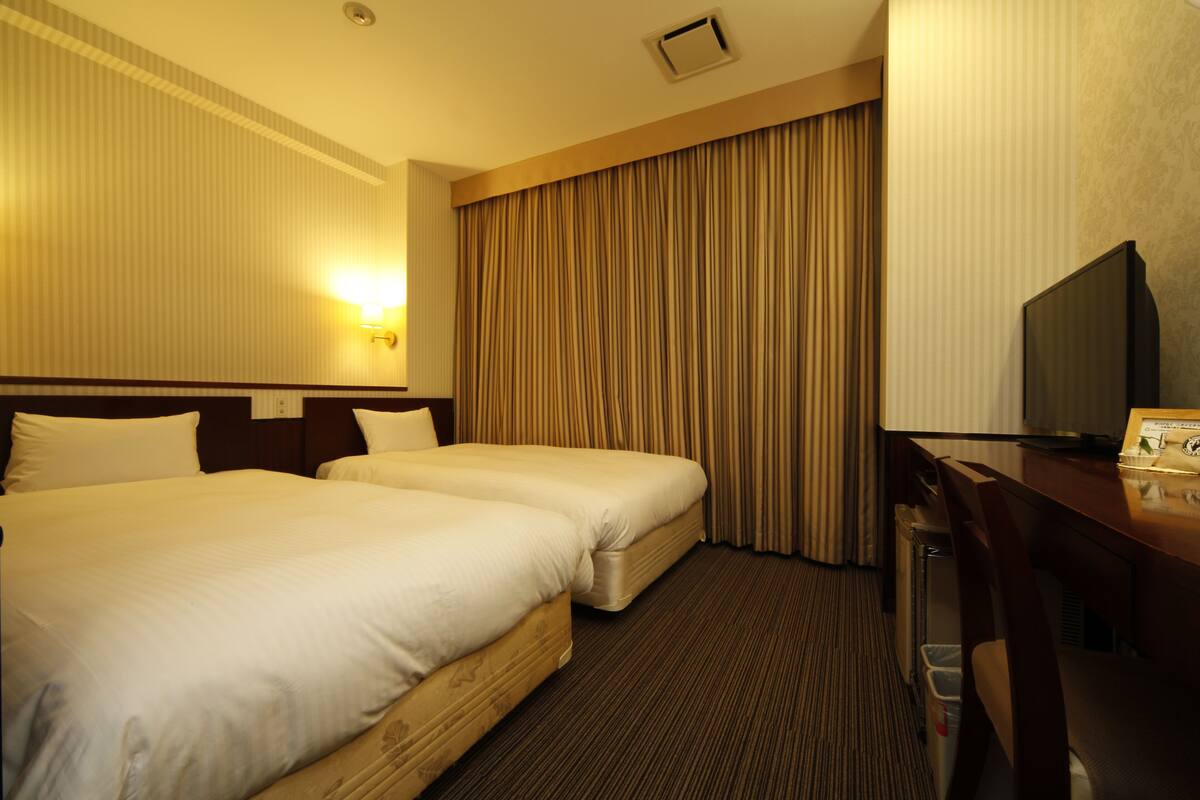 Ein Hotelzimmer mit zwei Betten, einem Schreibtisch, einem Fernseher und einem Vorhang.