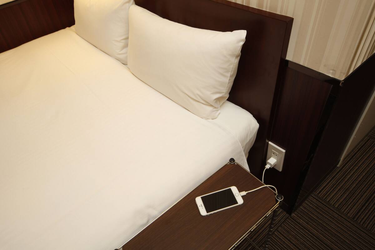 Ein Hotelbett mit weißen Laken, einem hölzernen Kopfteil und einem Nachttisch mit einem lädenden Handy.