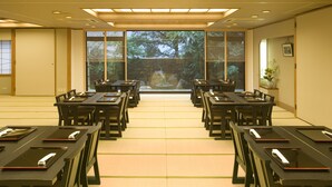 Un salón de conferencias con largas mesas y sillas, ventanas amplias con vistas a árboles y una estética japonesa tradicional.